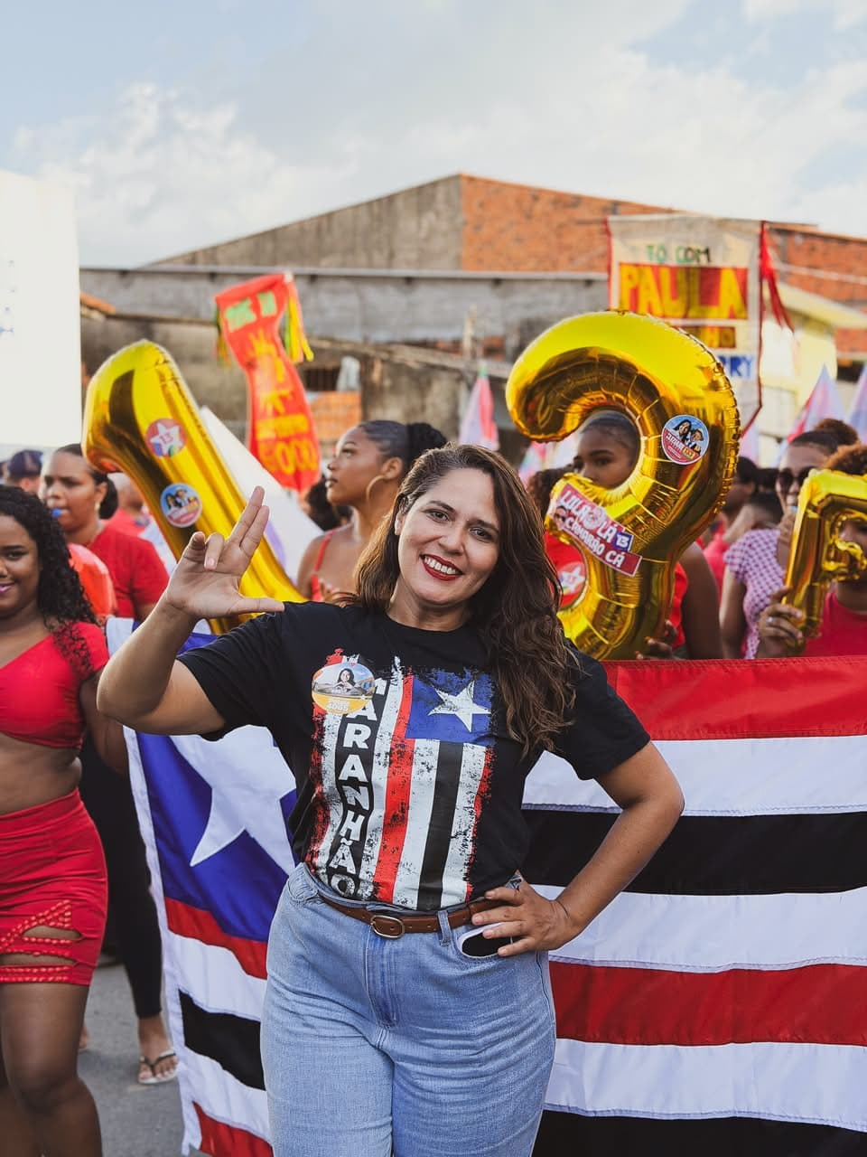 Pétala com o braço erguido em um ato de rua, à frente de uma bandeira do Maranhão, cercada por pessoas e balões dourados em forma de números.